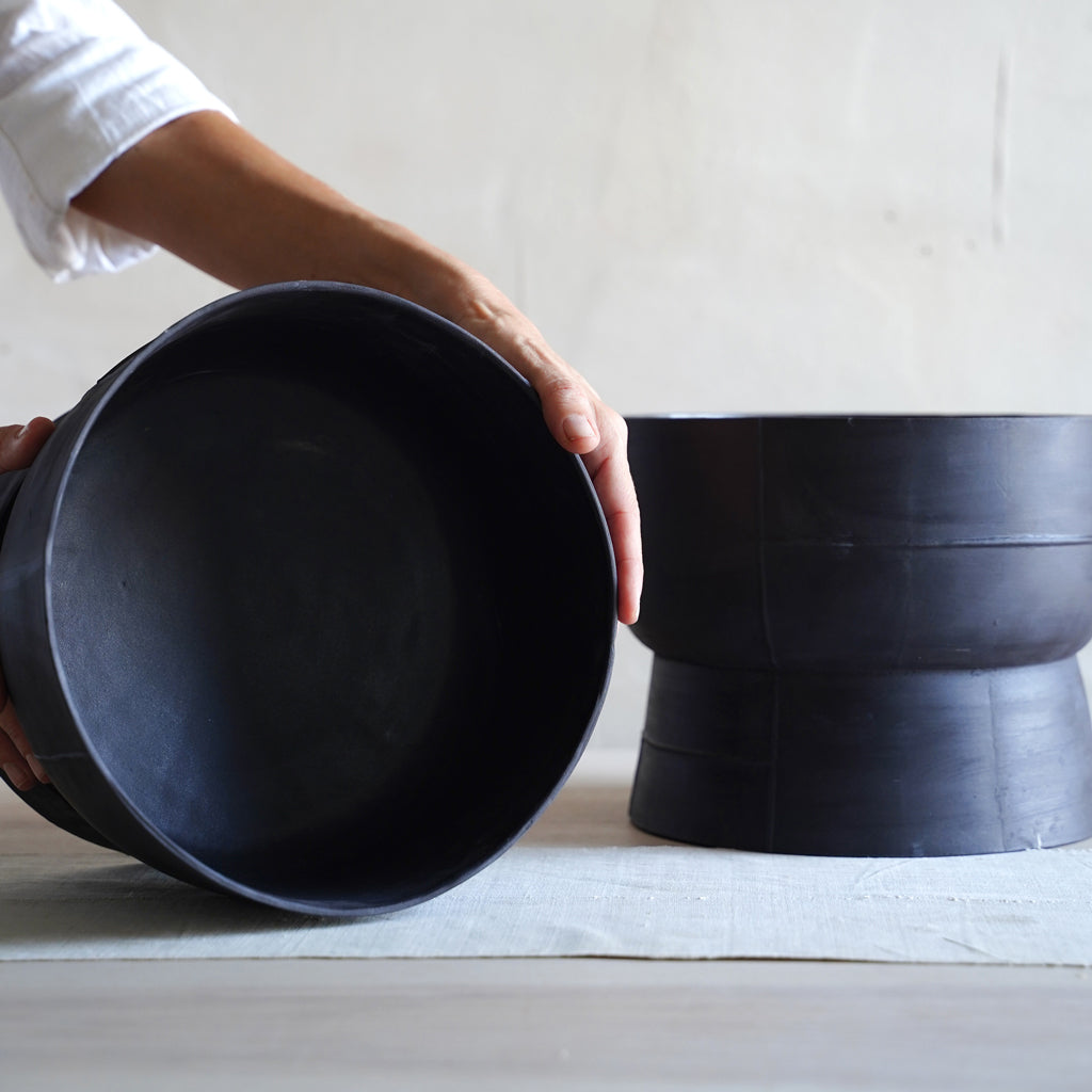 A person's hand holding a black stoneware bowl with a pedestal base, with another bowl placed on a linen surface in the background.