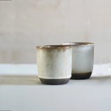 2 ivory cups photographed from the profile view. crafted from black stoneware and glazed a beige ivory. brown rim and an exposed base