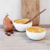 a stack of two porcelain coconut bowls with yellow interiors and a wooden spoon. Styled next to a single yellow coconut bowl, two pears and spices