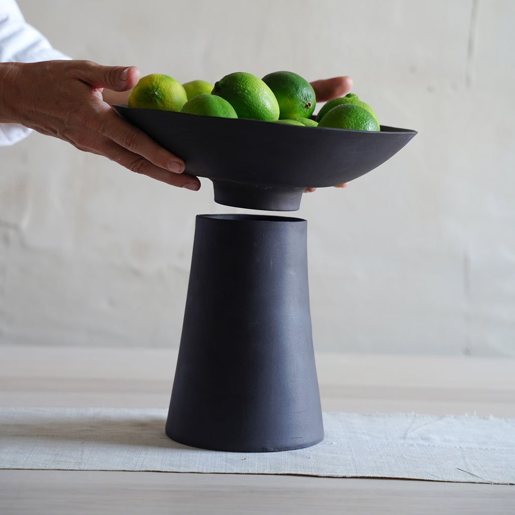 A black ceramic pedestal bowl with a vase-like base and a bowl on top filled with green limes, held by a person against a light background.