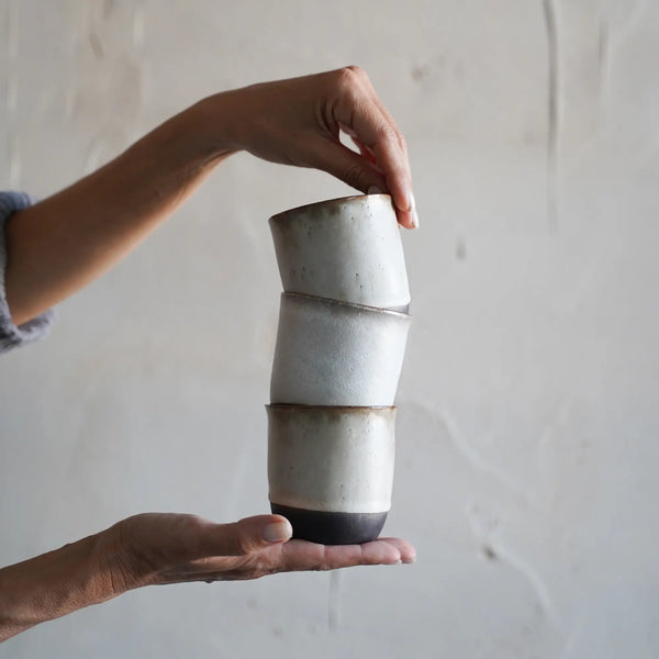 two hands artistically hold a stack of 3 handmade white ceramic cups with a light brown rim
