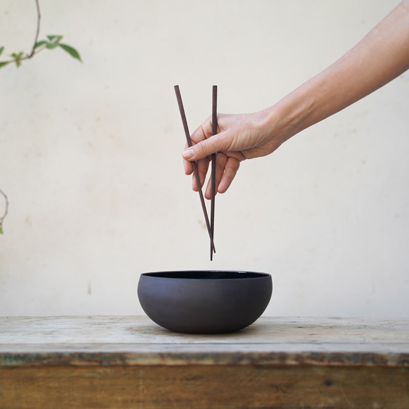 A hand holds chopsticks over a black ramen bowl, photographed from a profile view