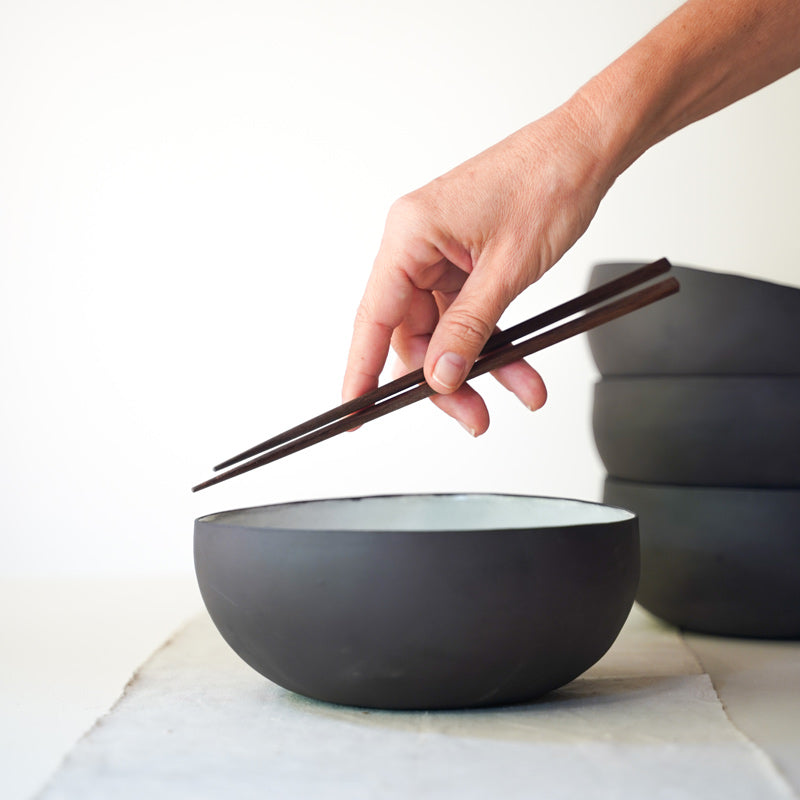 hand holds chopsticks over a black ramen bowl with a white interior