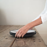 hands place a stack of four black and white stoneware dinner plates onto wooden table