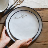 two hands set one medium white plate onto a large white plate. the plates have exposed black stoneware rims