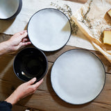 hand sets the table with two white plates and a black bowl. styled with cheese and flowers
