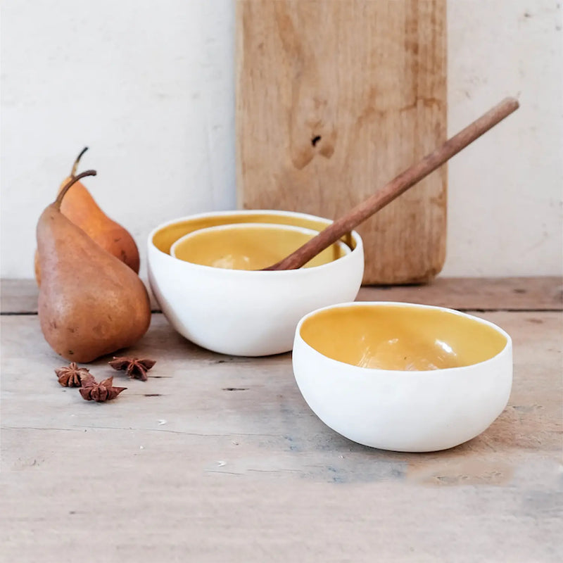 a stack of two porcelain coconut bowls with yellow interiors and a wooden spoon. Styled next to a single yellow coconut bowl, two pears and spices